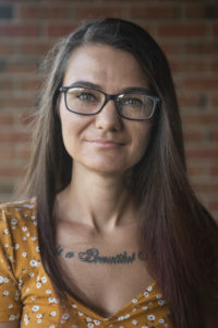 Headshot of Natalia Iwanek. Smiling white person and Toronto-based copy editor, proofreader, and writer with black framed glasses, long brown hair, and yellow shirt against a blurred, brown brick background.